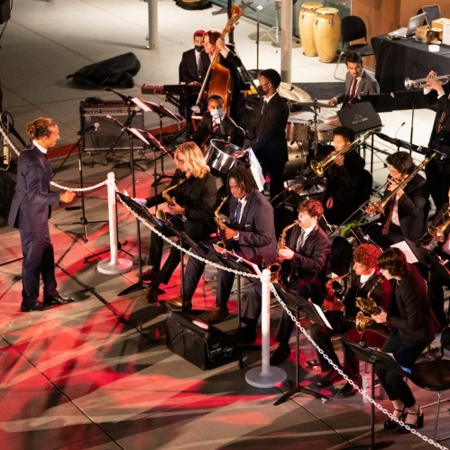 A jazz band performs outdoors, featuring musicians with various instruments and a conductor in a suit.