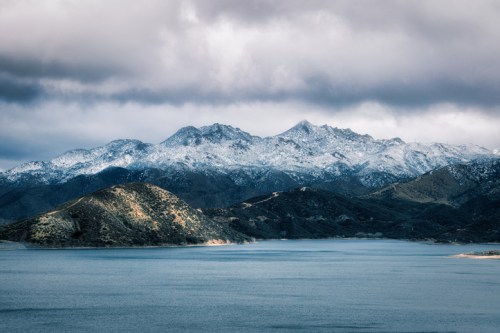 Snow-capped mountains rise above a calm lake, under a cloudy sky, with rolling hills in the foreground.