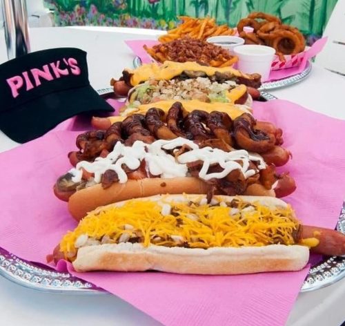 A colorful platter of hot dogs topped with various ingredients, served with fries and pretzels, alongside a pink cap.