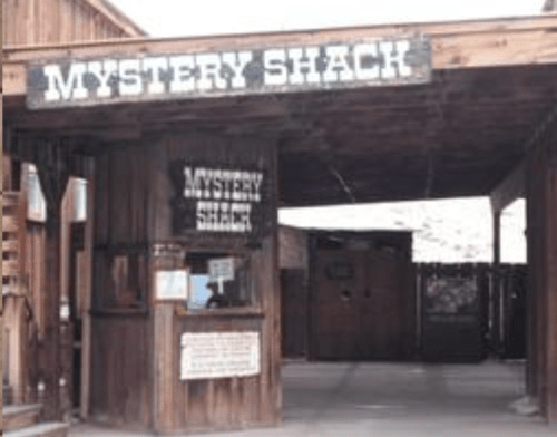 Entrance to the Mystery Shack, featuring wooden structures and signs, set in a rustic outdoor environment.