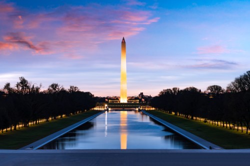 The Washington Monument illuminated at dusk, reflecting in the nearby pool, surrounded by trees and a colorful sky.