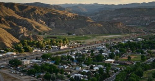 Aerial view of a small town nestled between mountains, with a train running alongside and greenery in the valley.