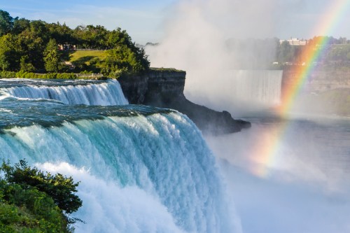 A vibrant waterfall cascades into a misty river, with a rainbow arcing above the scene and lush greenery nearby.