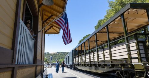 A sunny day at a train station with an American flag, a train car, and people walking along the platform.