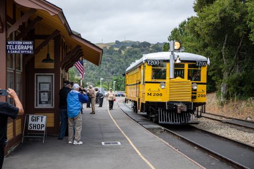 A vintage yellow train at a station with people waiting and a telegraph office nearby.