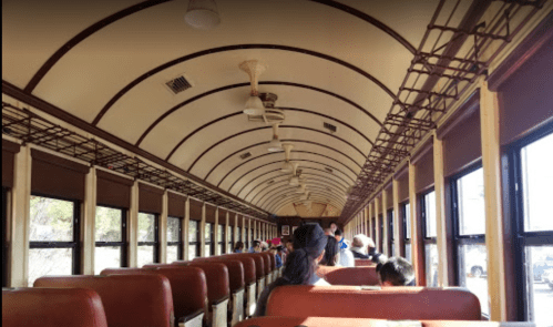 Interior of a vintage train car with rows of brown seats and large windows, showing passengers seated inside.