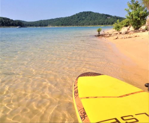 A yellow paddleboard rests on a sandy shore by a calm lake, surrounded by green hills and clear blue water.
