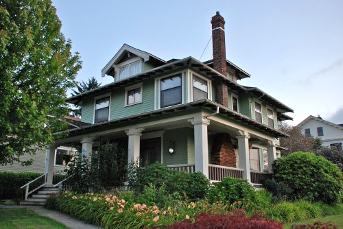 A charming green house with a porch, surrounded by lush gardens and trees, under a clear sky.