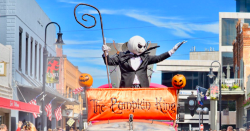 A character in a black suit and white face stands on a float, waving, with pumpkins and a "Pumpkin King" banner.