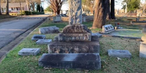 A weathered gravestone with the name "Gwyant" surrounded by fallen stones in a cemetery. Trees and a road are visible.