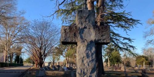 A weathered stone cross in a cemetery, surrounded by trees and gravestones under a clear blue sky.