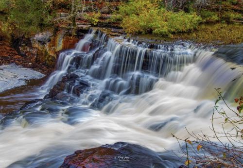A serene waterfall cascades over rocks, surrounded by lush greenery and autumn foliage.