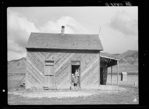 A wooden house with a slanted roof, a woman and child at the door, and a dog outside, set against a mountainous backdrop.