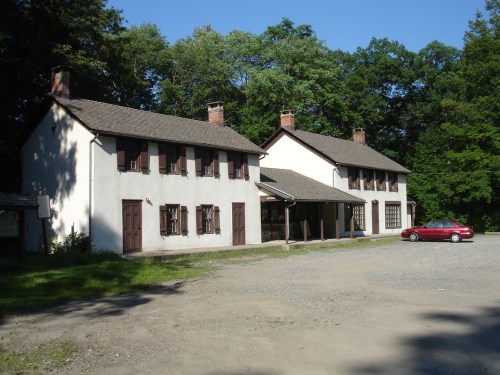 Two rustic buildings with brown shutters, surrounded by greenery, and a red car parked in the gravel lot.