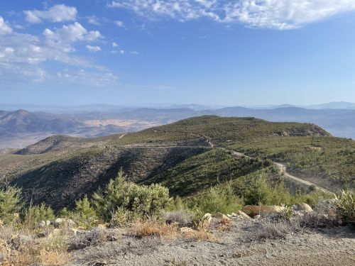 A scenic view of rolling hills and mountains under a blue sky, with a winding road visible in the foreground.