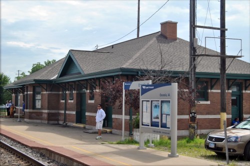 A train station with a brick building, platform, and a sign for Cassella Ave. A person stands nearby.