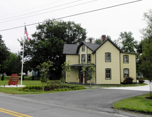 A yellow two-story house with a flagpole and sign in front, surrounded by trees and a well-maintained lawn.