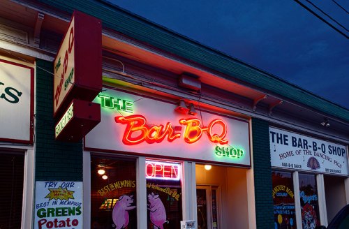 Neon sign of "The Bar-B-Q Shop" glowing at dusk, with an "Open" sign visible in the window.