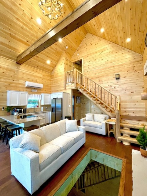 Cozy wooden cabin interior with high ceilings, a staircase, and a living area featuring a sofa and a glass coffee table.