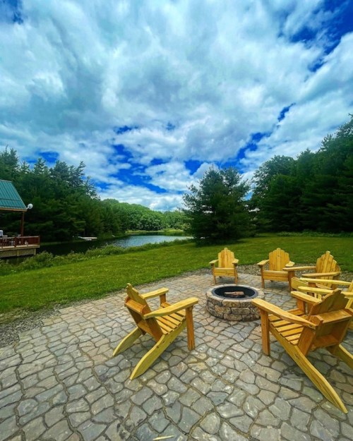 A serene outdoor scene featuring wooden chairs around a fire pit, with a pond and trees under a cloudy sky.