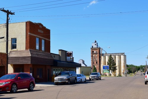 A small town street featuring brick buildings, parked cars, and grain silos under a clear blue sky.