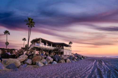 A beachside restaurant at sunset, surrounded by palm trees and rocky shoreline under a colorful sky.