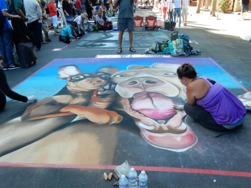 A street artist creates a colorful chalk drawing of dogs on pavement, surrounded by people at an outdoor event.