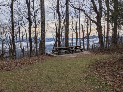 A picnic table surrounded by trees overlooks a misty valley and distant mountains under a cloudy sky.