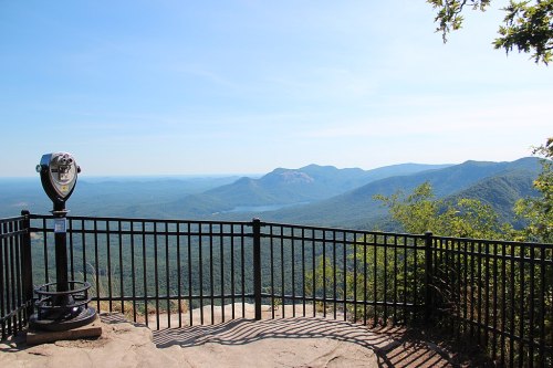 A scenic overlook with a viewing telescope, surrounded by a black fence, showcasing distant mountains and a clear blue sky.