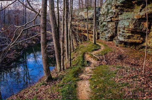 A winding trail beside a river, flanked by rocky cliffs and bare trees, with autumn leaves scattered on the ground.