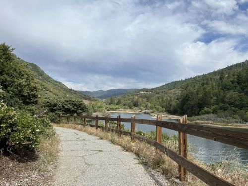 A winding path beside a river, surrounded by lush greenery and mountains under a partly cloudy sky.