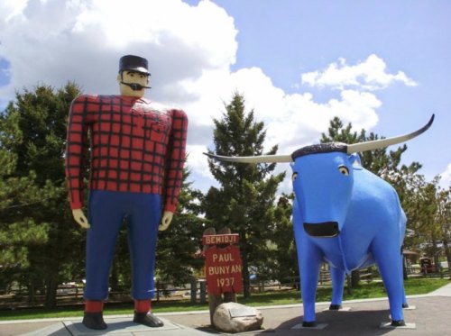 A large statue of Paul Bunyan in a plaid shirt stands next to a blue ox, with trees and clouds in the background.