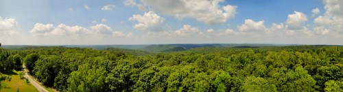 A panoramic view of lush green forests and rolling hills under a bright blue sky with fluffy white clouds.