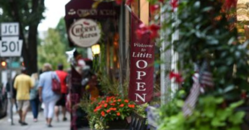 A charming street scene in Lititz with an "Open" sign and colorful flowers, as people stroll by.