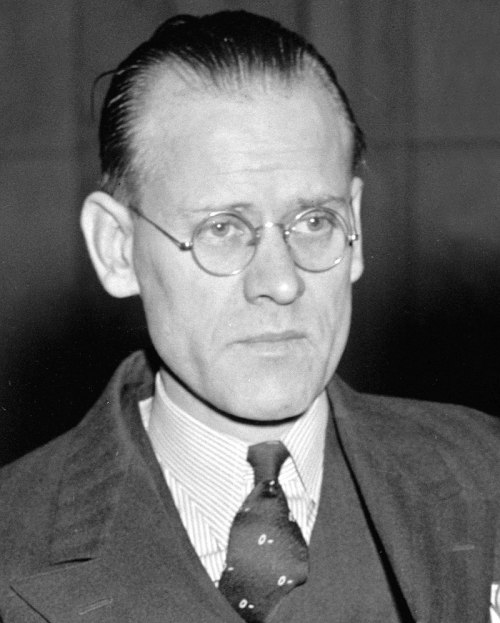 A serious man with glasses and slicked-back hair, wearing a suit and tie, looking directly at the camera.