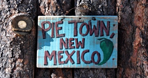 A handmade sign saying "Pie Town, New Mexico" hangs on a log fence.