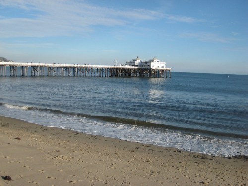 A wooden pier extends over calm blue waters, with a sandy beach in the foreground and a clear sky above.