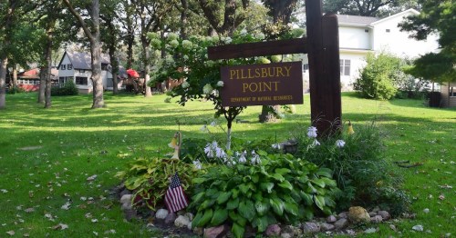 Sign for Pillsbury Point, surrounded by greenery and flowers, with a small American flag nearby.