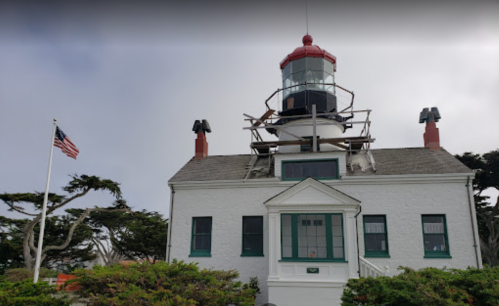 A lighthouse with a red and white tower beside a white building, surrounded by trees and an American flag.