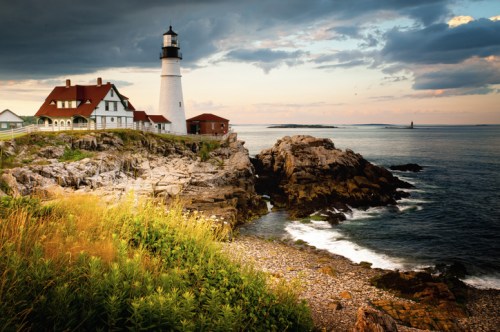 A scenic lighthouse on rocky shores, surrounded by grass and ocean waves under a cloudy sky at sunset.