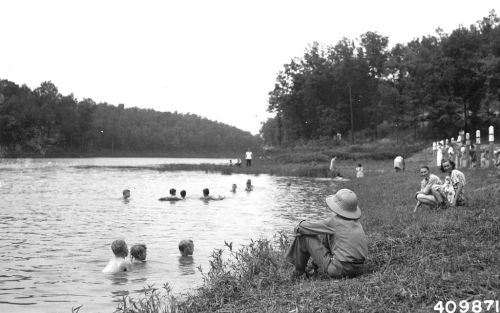 A black-and-white photo of people swimming and relaxing by a river, surrounded by trees and grassy banks.