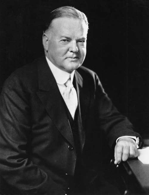A formal portrait of a man in a suit, seated with a serious expression, against a dark background.