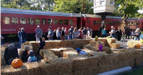 A crowd gathers around a hay maze with pumpkins, near a vintage red train in a festive outdoor setting.