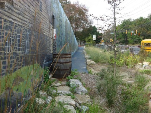 A pathway beside a mural, featuring a barrel and overgrown grass, with a road and school bus in the background.