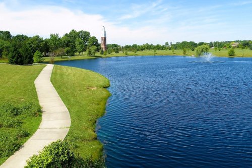 A serene park scene featuring a pond, walking path, green grass, and a distant clock tower under a clear blue sky.
