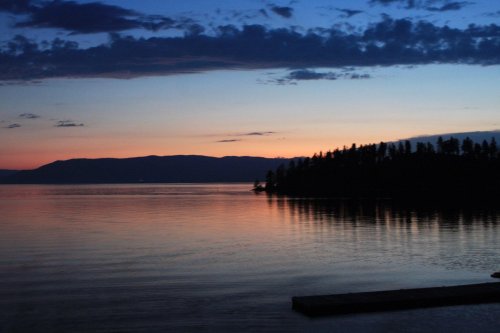 A serene sunset over a calm lake, with silhouettes of trees and mountains reflecting on the water's surface.