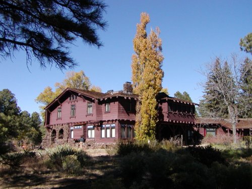 A large, rustic red house surrounded by trees and greenery under a clear blue sky.