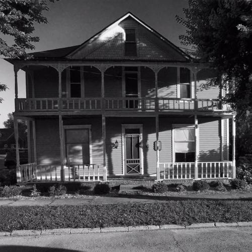 A vintage two-story house with a porch, surrounded by greenery, captured in black and white.