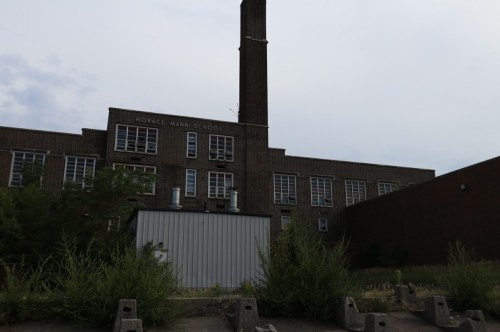 Abandoned Horace Mann School building with a tall chimney, surrounded by overgrown vegetation and a gray sky.
