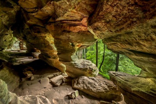 A rocky cave interior with colorful stone formations and a view of lush green trees outside.